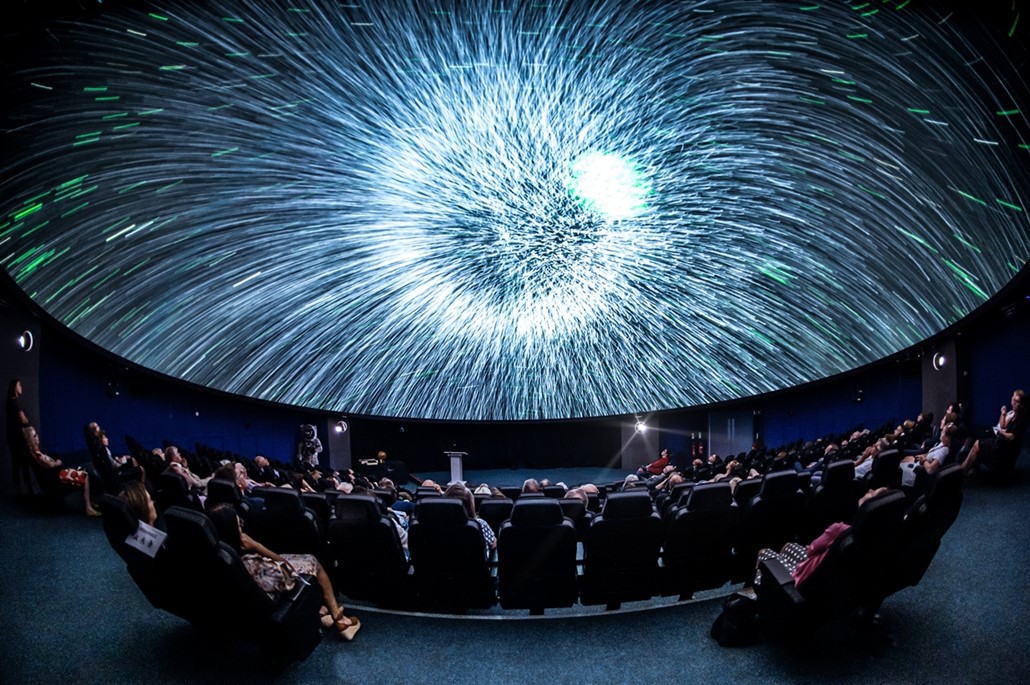 article thumb - A photo of inside the Planetarium. A bright white shining light on the curved dome with an audience watching.